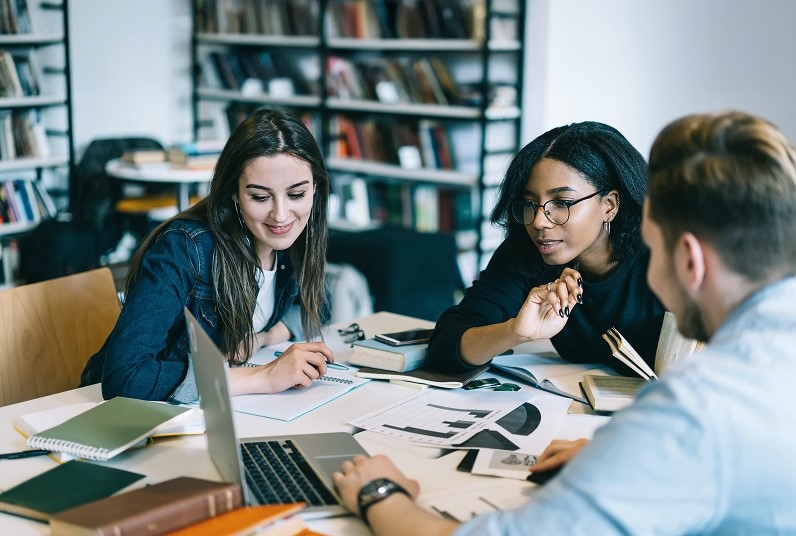 Drei junge Menschen sitzen an einem Tisch mit Lernmaterial und schauen interessiert auf einen Laptop